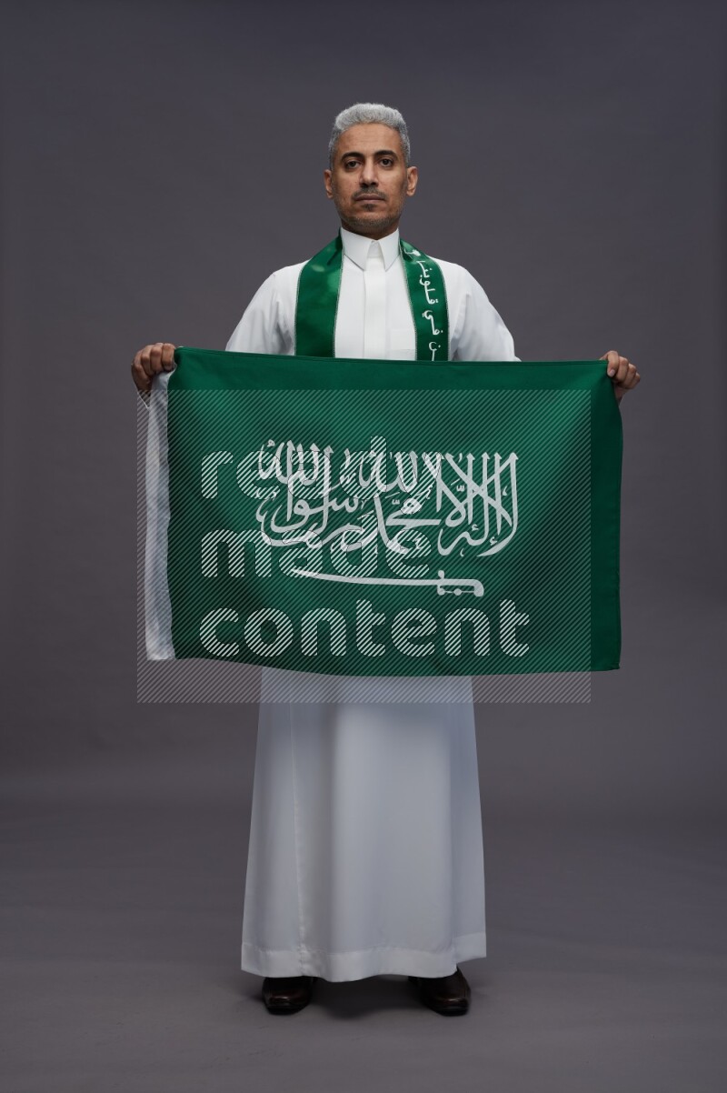 Saudi man wearing thob standing holding Saudi flag on gray background