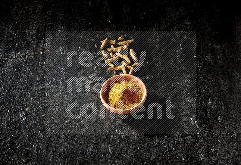 A wooden bowl full of turmeric powder with dried turmeric fingers on textured black flooring