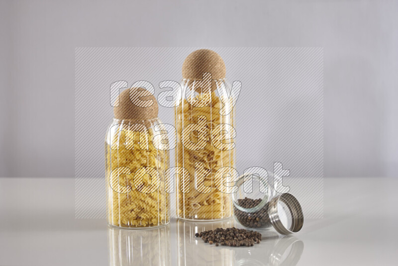 Raw pasta in glass jars with black peppers on light grey background