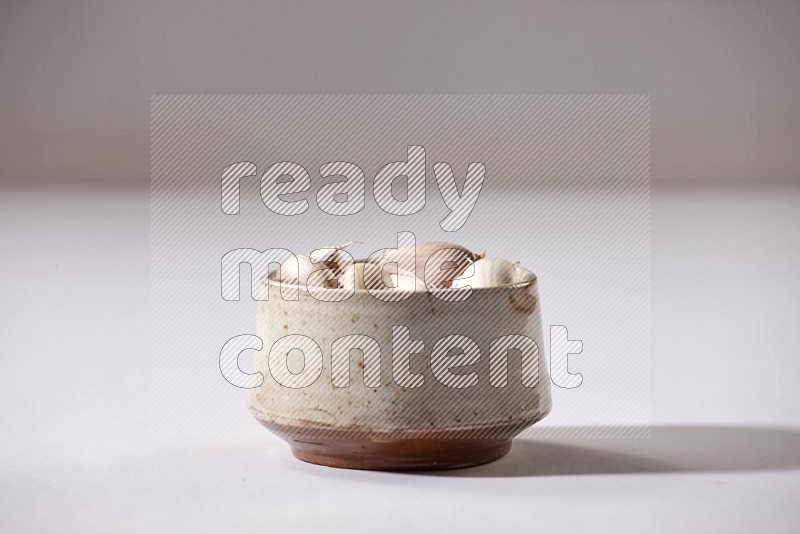 A beige pottery bowl full of garlic cloves on a white flooring in different angles