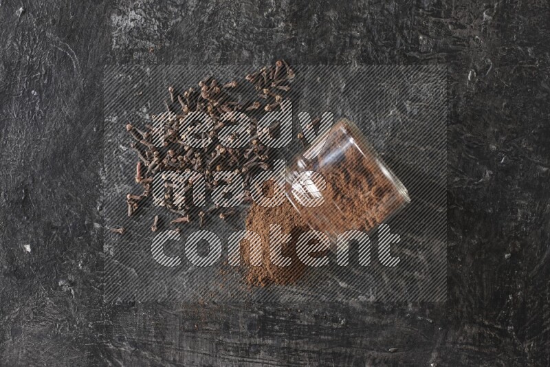 A flipped glass jar full of cloves powder with cloves spread on a textured black flooring