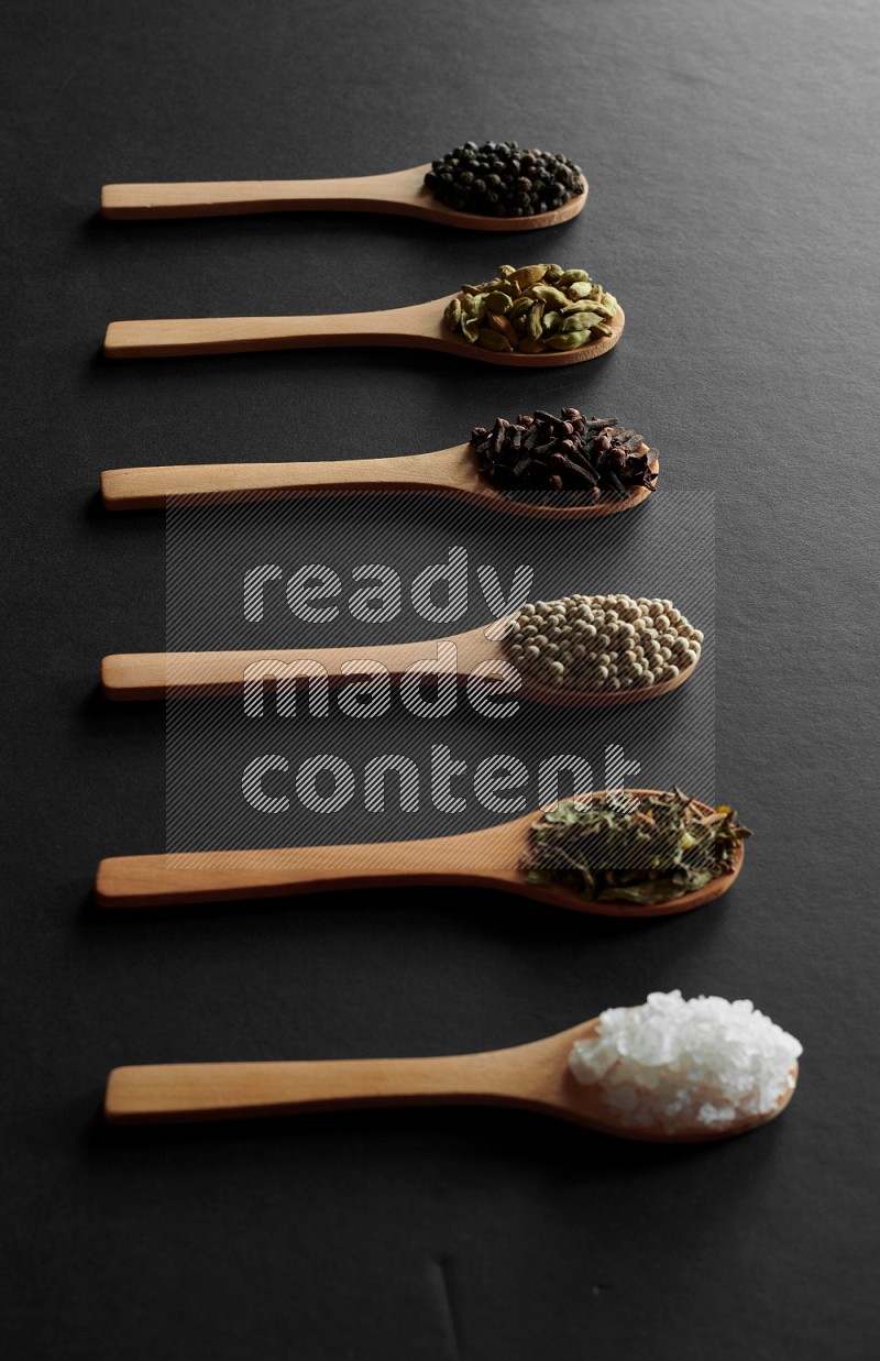 wooden spoons filled with white peppers, cloves, cardamom, salt, black peppers and basil on black flooring