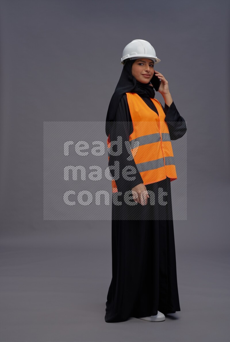 Saudi woman wearing Abaya with engineer vest standing talking on phone on gray background