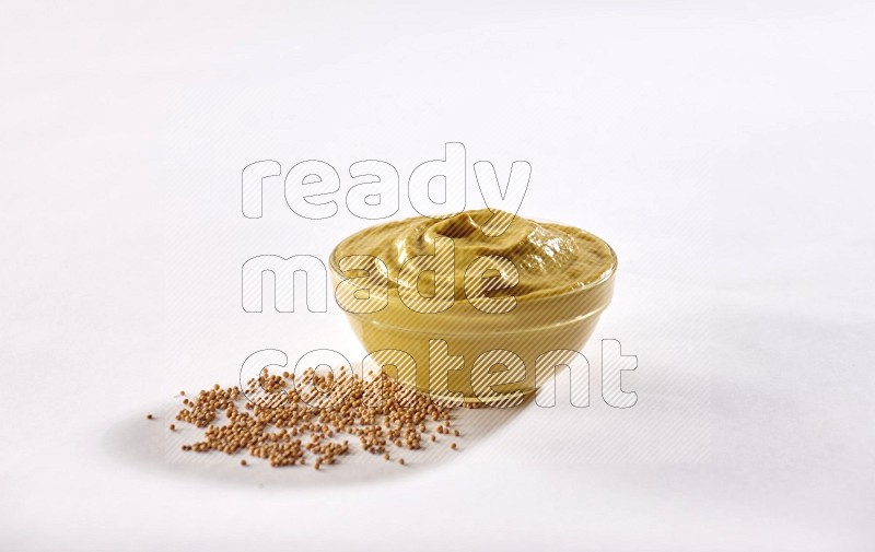 A glass bowl full of mustard paste with mustard seeds underneath on white flooring
