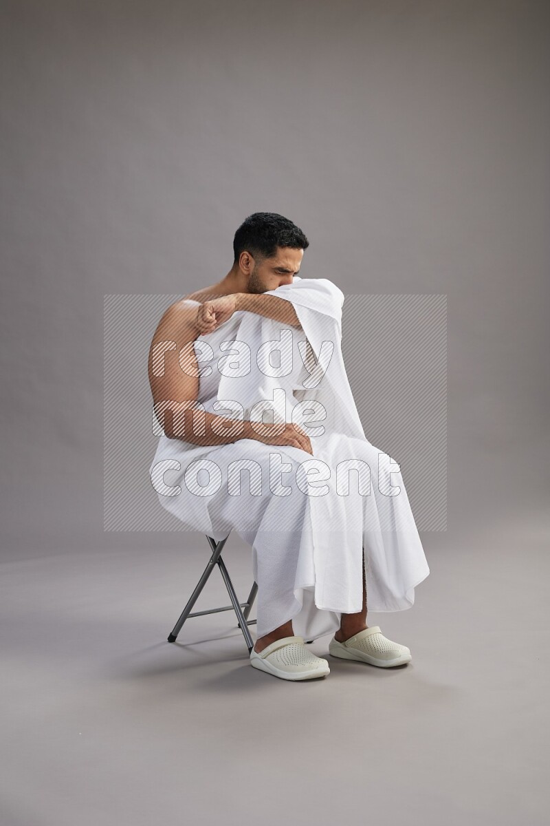 A man wearing Ehram sitting on chair showing how to sneeze on gray background