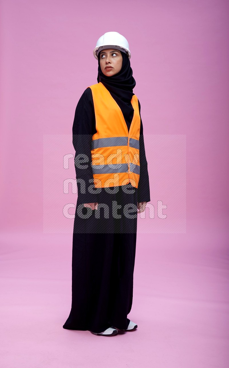 Saudi woman wearing Abaya with engineer vest and helmet standing interacting with the camera on pink background