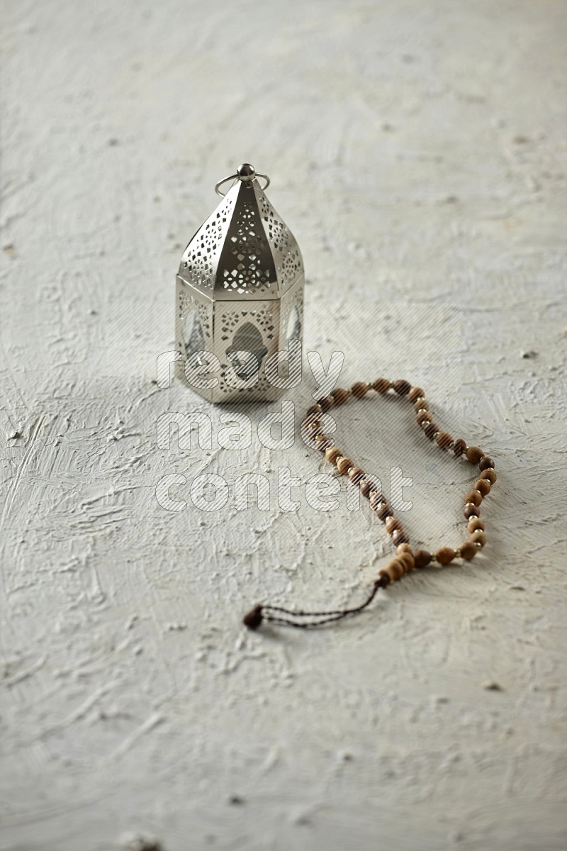 A silver lantern with different drinks, dates, nuts, prayer beads and quran on textured white background