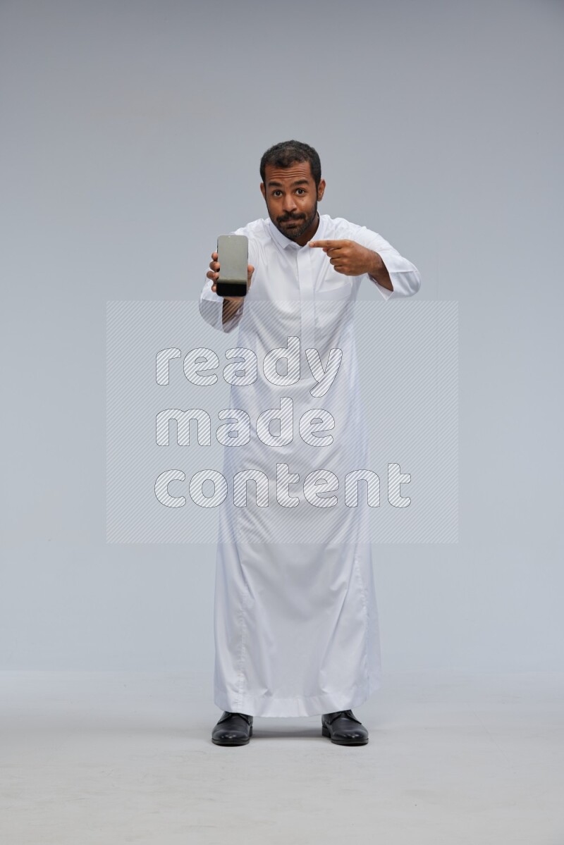 Saudi man wearing Thob standing showing phone to camera on Gray background