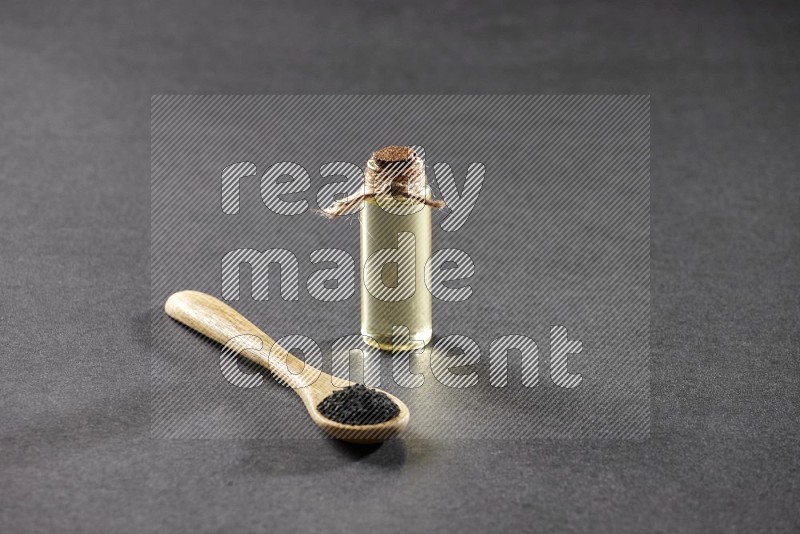 A wooden spoon full of black seeds and a glass bottle of black seeds oil on a black flooring
