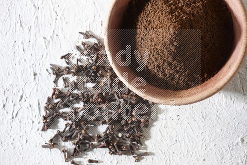A wooden bowl full of cloves powder with whole cloves beside it on a textured white flooring