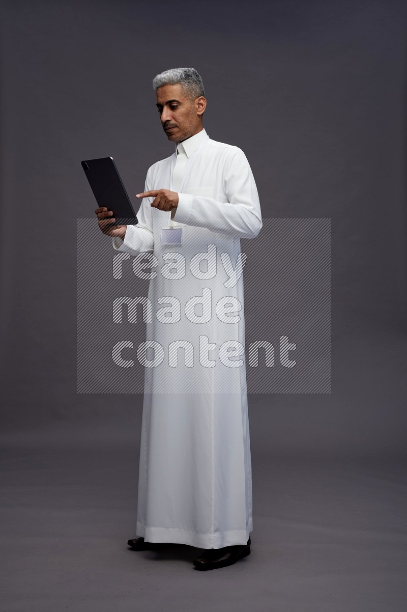 Saudi man wearing thob with neck strap employee badge standing working on tablet on gray background