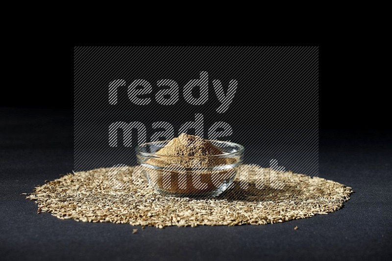 A glass bowl full of cumin powder surrounded by cumin seeds on black flooring