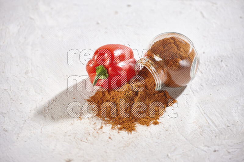 A glass jar full of ground paprika powder flipped with some spilling powder on white background