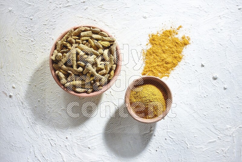 2 wooden bowls, one full of turmeric powder and the other full of dried turmeric whole fingers on a textured white flooring