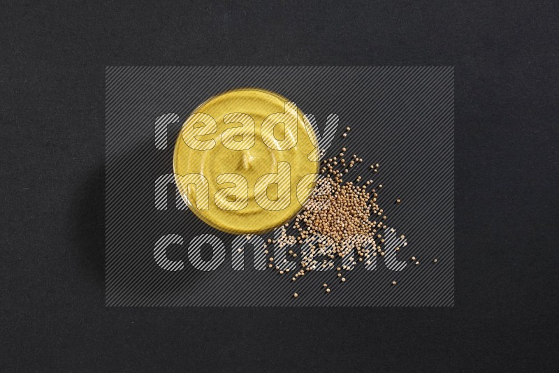 A glass bowl full of mustard paste with mustard seeds underneath on black flooring