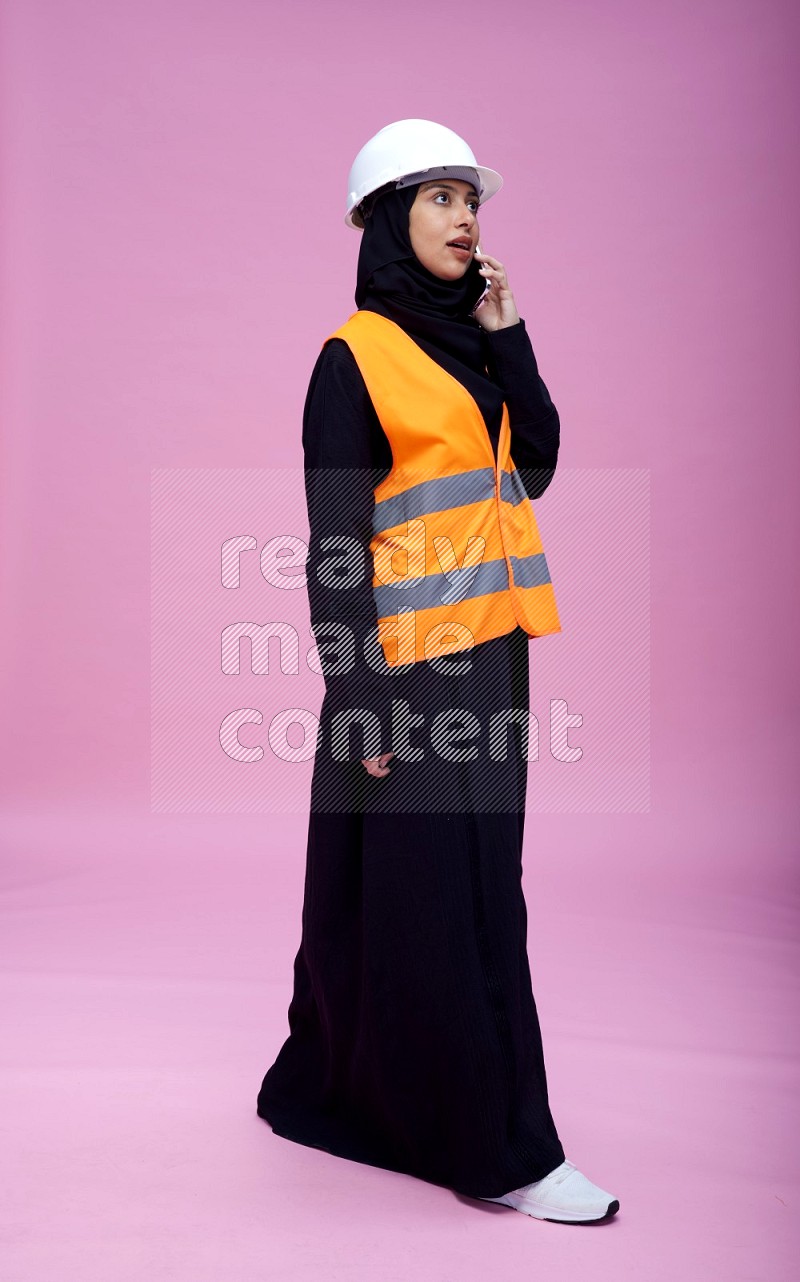 Saudi woman wearing Abaya with engineer vest and helmet standing talking on phone on pink background