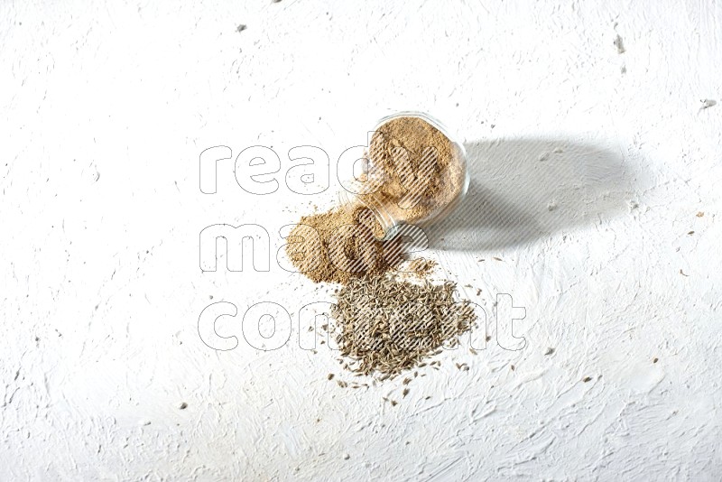 A flipped glass spice jar full of cumin powder and powder spilled out with cumin seeds on textured white flooring