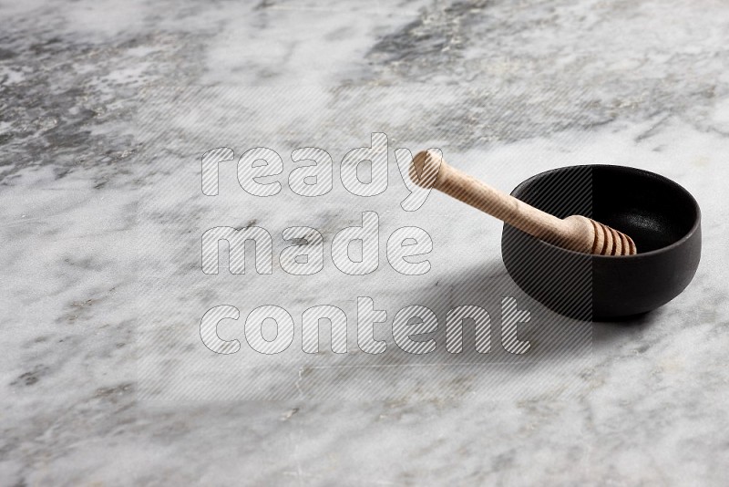Black Pottery bowl with wooden honey handle in it, on grey marble flooring, 45 degree angle