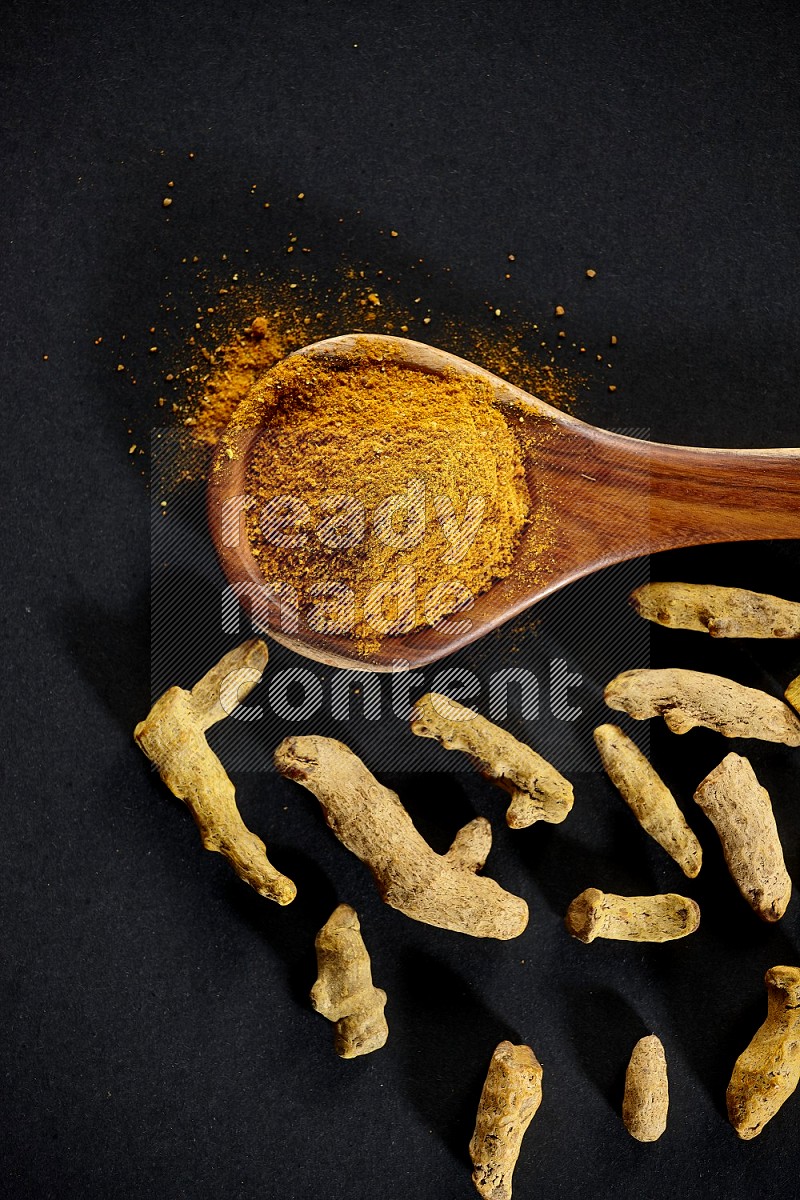 A wooden ladle full of turmeric powder with dried turmeric fingers on black flooring