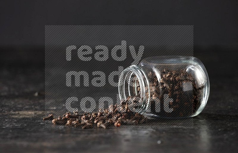 A flipped glass spice jar full of cloves and cloves came out of it on textured black flooring