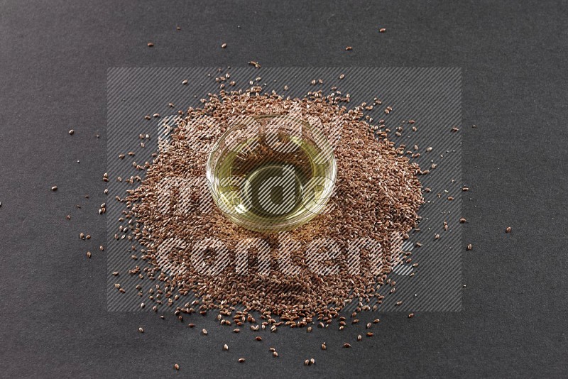 A glass bowl full of flaxseeds oil surrounded by the seeds on a black flooring