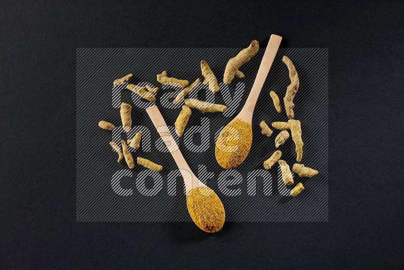 2 wooden spoons full of turmeric powder with dried turmeric fingers on black flooring