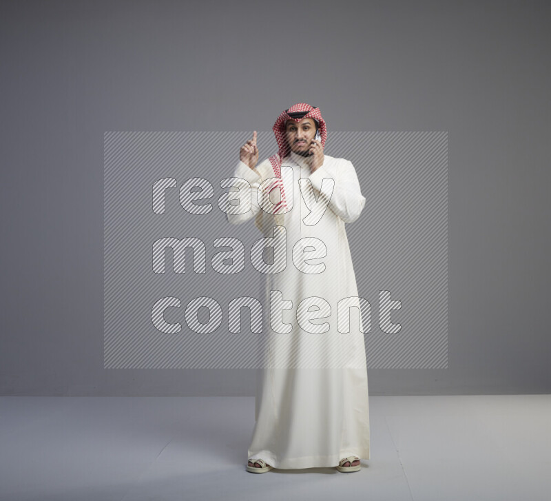 A Saudi man standing wearing thob and red shomag talking on phone on gray background