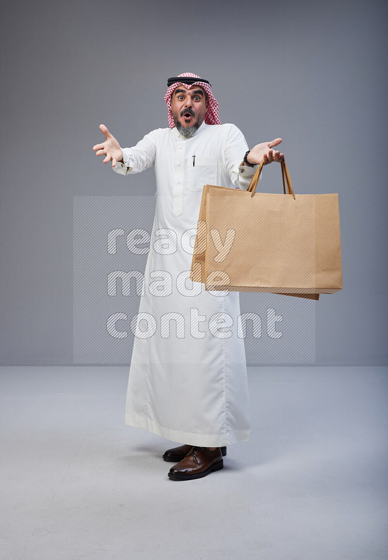 Saudi man Wearing Thob and red Shomag standing holding shopping bag on Gray background