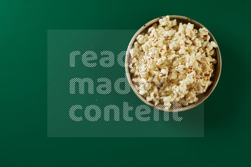 A beige ceramic bowl full of popcorn on a green background in a top view shot
