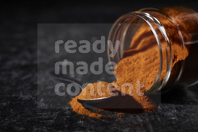 A glass jar full of ground paprika powder flipped with some spilling powder on black background