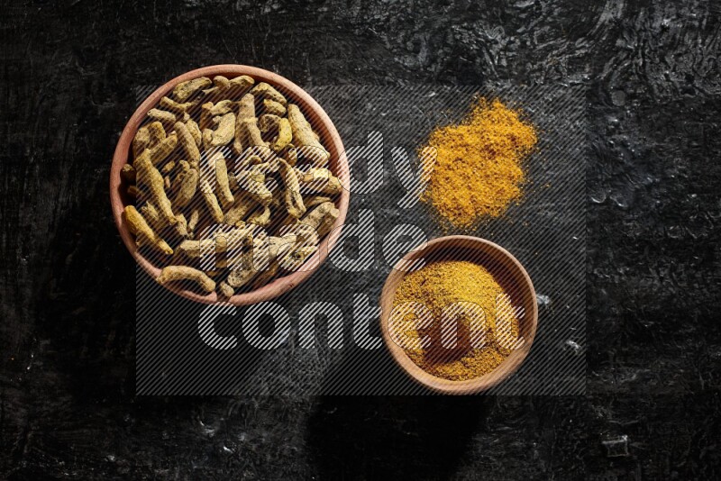 2 wooden bowls, one full of turmeric powder and the other full of dried turmeric whole fingers on a textured black flooring