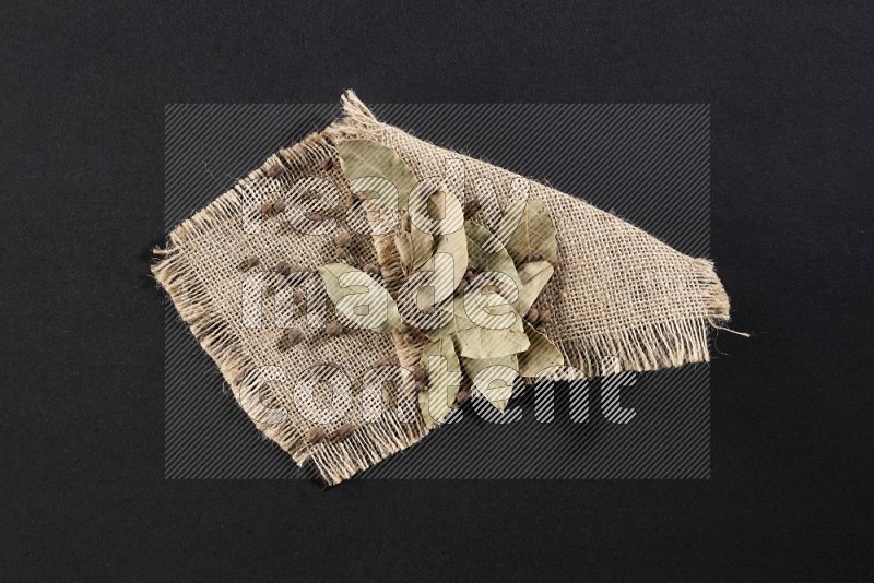 Dried bay leaves and allspice berries on a piece of burlap on black flooring