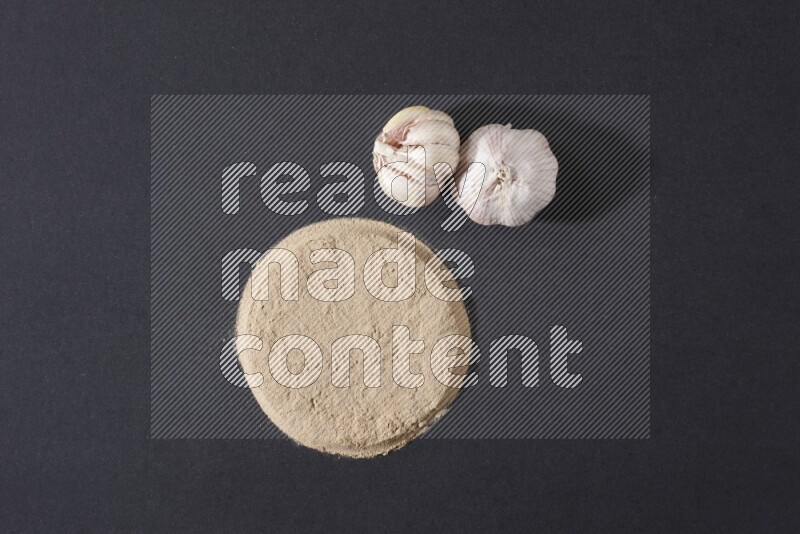 ثوم بودرة على شكل دائرة وبجانبه 2 رؤوس ثوم على خلفية سوداء