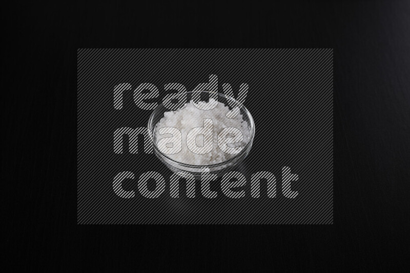 A glass bowl full of coarse sea salt crystals on black background