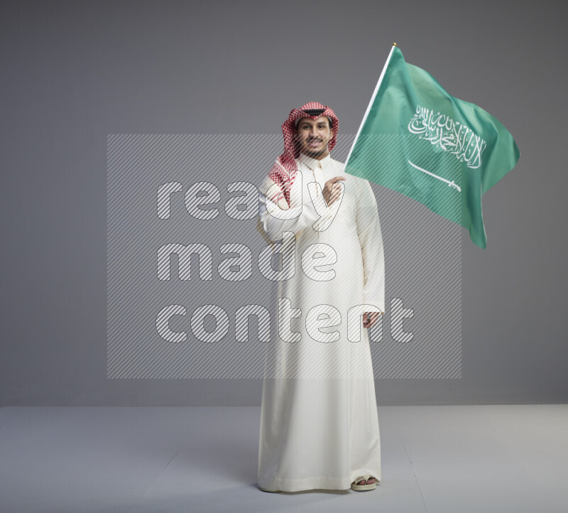 A Saudi man standing wearing thob and red shomag raising big Saudi flag on gray background