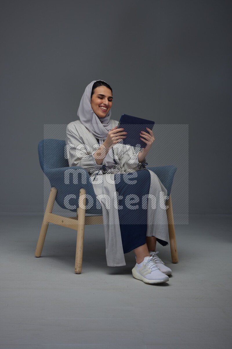 A Saudi female wearing a light gray Abaya and head scarf sitting on a dark grey chair while holding her iPad on a grey background