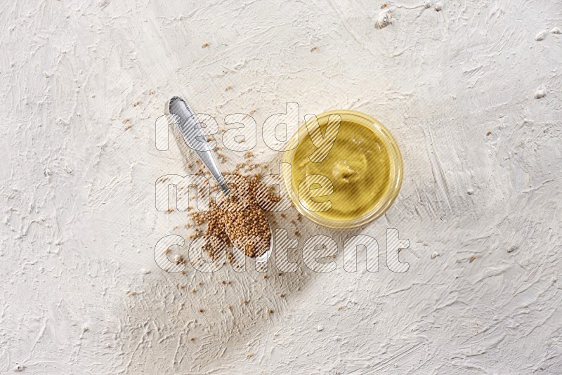 A glass jar full of mustard paste and a metal spoon full of mustard seeds on a textured white flooring