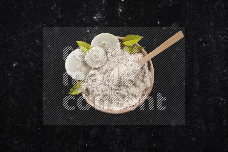 A wooden bowl full of onion powder with a wooden spoon in it with some sliced onions on black background