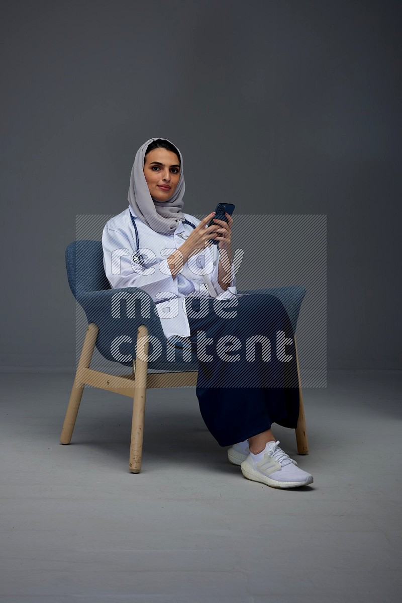 A doctor wearing a light gray head scarf sitting on a dark grey chair and using a laptop while talking in the phone eye level on a grey background