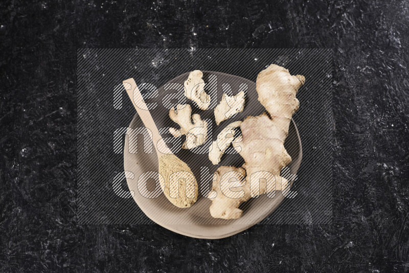 A wooden spoon full of ground ginger powder with fresh and dried ginger, all on a pottery plate on black background