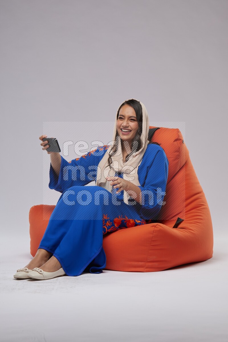 A Saudi woman wearing jalabiya sitting on an orange beanbag and taking selfie