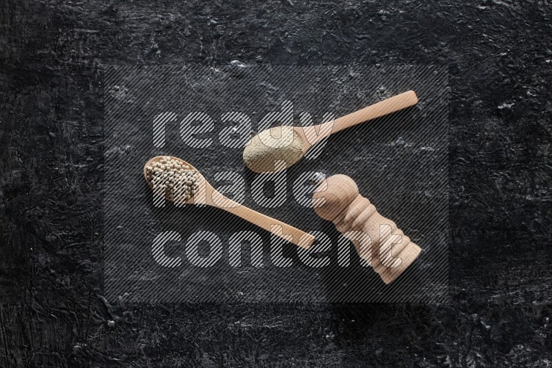 2 wooden spoons one full of white pepper powder and the other with pepper beads and a wooden pepper mill on textured black flooring