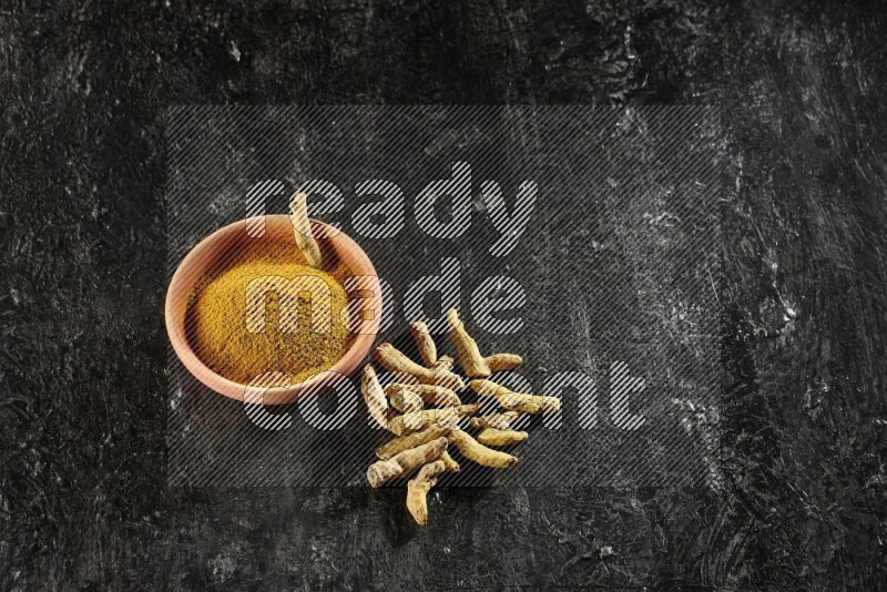 A wooden bowl full of turmeric powder with dried turmeric whole fingers on textured black flooring