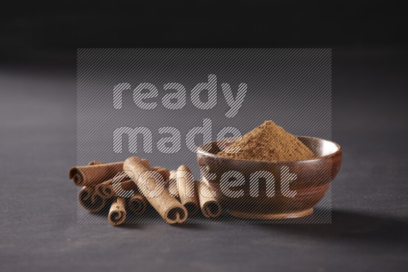 Cinnamon sticks stacked beside a wooden bowl full of cinnamon powder on black background