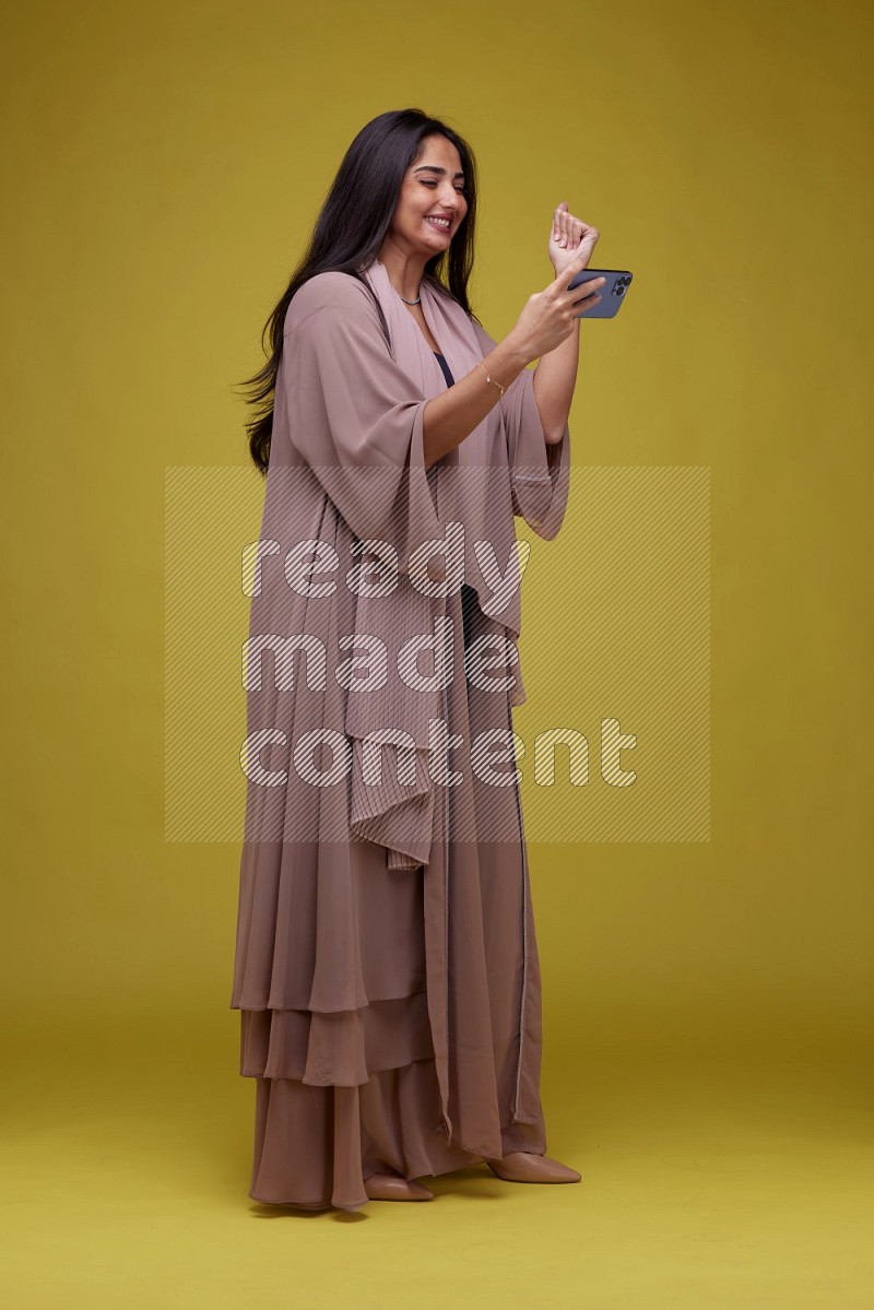 A woman Playing a Game on her smartphone on a Yellow Background wearing Brown Abaya