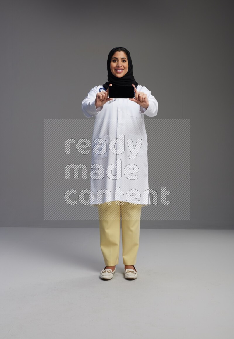 Saudi woman wearing lab coat with stethoscope standing showing phone to camera with sign in the back on Gray background