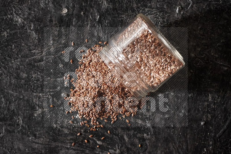 A glass jar full of flaxseeds flipped and seeds spread out on a textured black flooring
