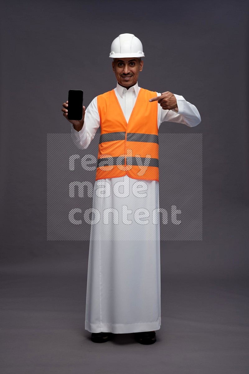 Saudi man wearing thob with engineer vest standing showing phone to camera on gray background