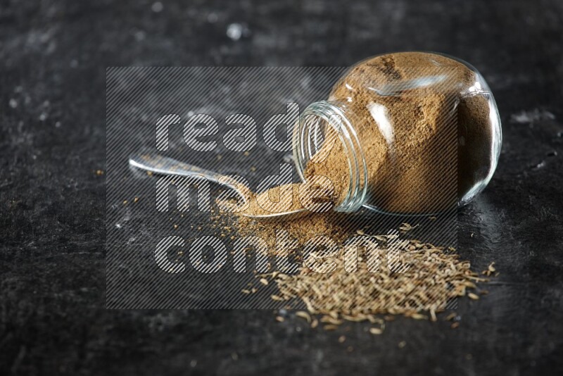 A flipped glass spice jar and a metal spoon full of cumin powder and powder spilled out with cumin seeds on a textured black flooring