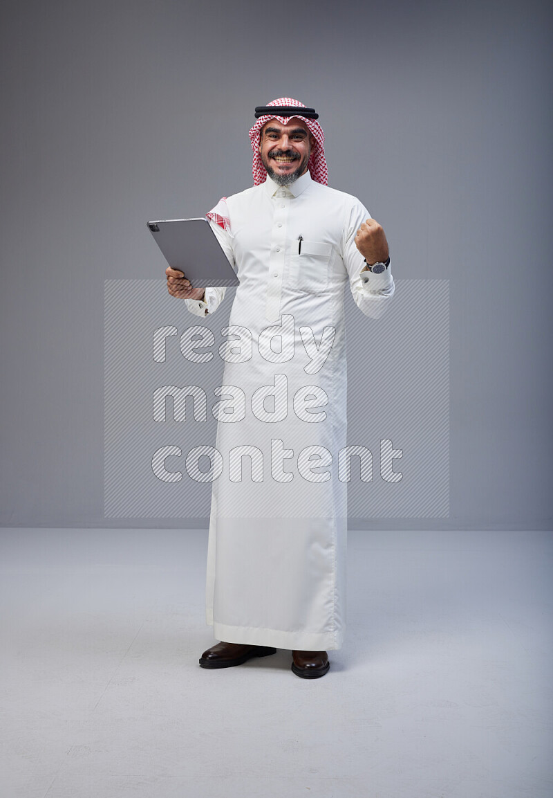 Saudi man Wearing Thob and red Shomag standing working on tablet on Gray background